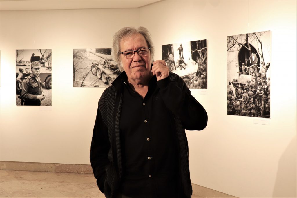 Sérgio Godinho, cantor, compositor, letrista e escritor, em primeiro plano, vestido de preto. Atrás, paredes adornadas com fotografias a preto e branco.