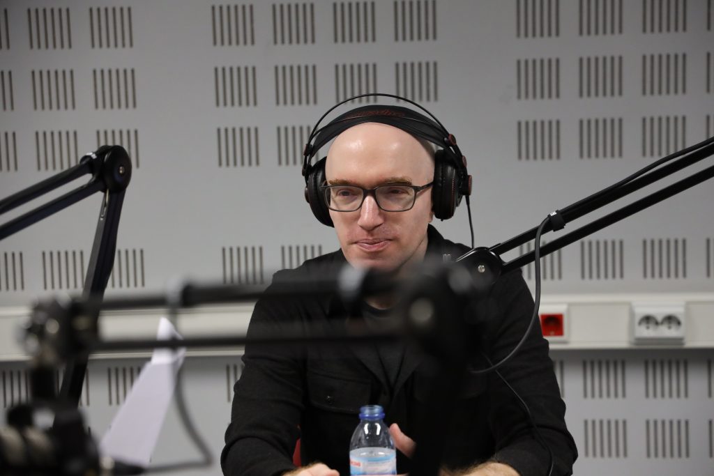 Daniel Gallant está num estúdio de rádio, com auscultadores e em frente a um microfone. É careca, usa óculos e veste uma camisa negra.