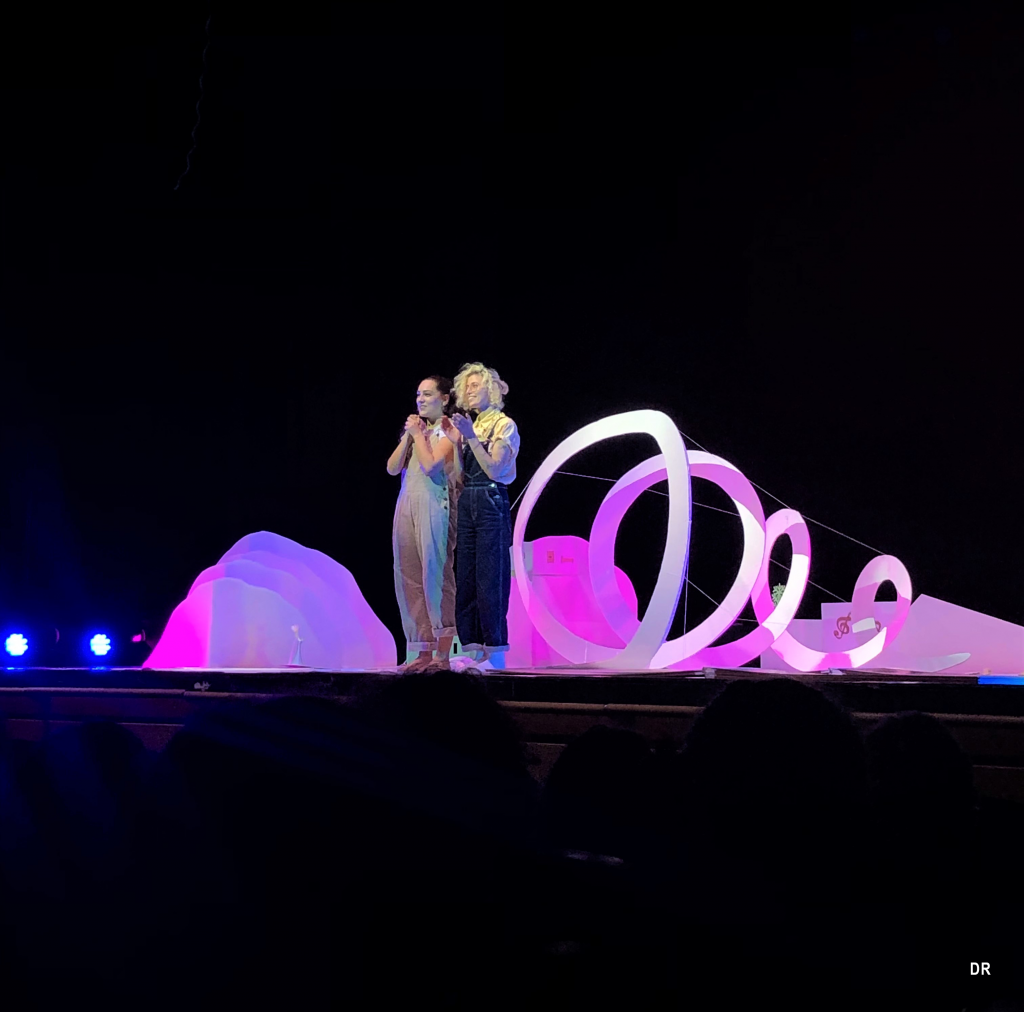 Duas artistas num palco, iluminadas, agradecem ao público. O resto do cenário está a preto à exceção de elementos circulares e arredondados de cores roxo e rosa.