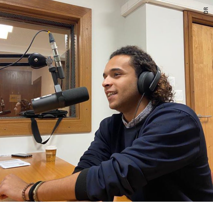 Jovem estudante de comunicação fala ao microfone num estúdio de rádio. Tem auscultadores, cabelo comprido e veste uma camisola azul escura.