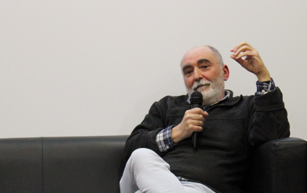 O cartoonista Luís Afonso posa está sentado num sofá preto, a olhar em frente e a falar para um microfone. Tem barba e cabelo branco e usa um pólo preto e uma camisa ao xadrez azul e branca.