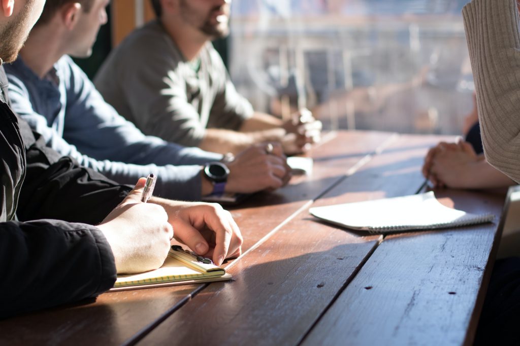 Grupo de pessoas reunidas numa mesa. Em cima da mesa, blocos de notas onde fazem apontamentos. Uma das pessoas do grupo fala e as outras olham para ela.