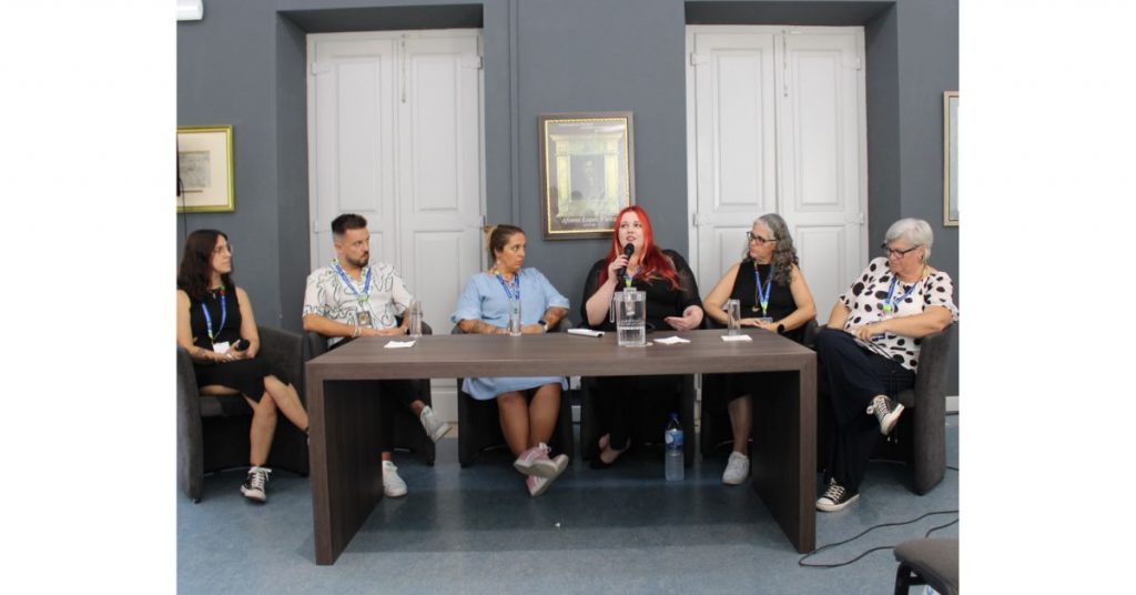 Conjunto de seis participantes do encontro Entre Palavras sentados a uma mesa. O grupo é composto por cinco mulheres e um homem. Uma das mulheres, ao centro, fala para um microfone que tem na mão.