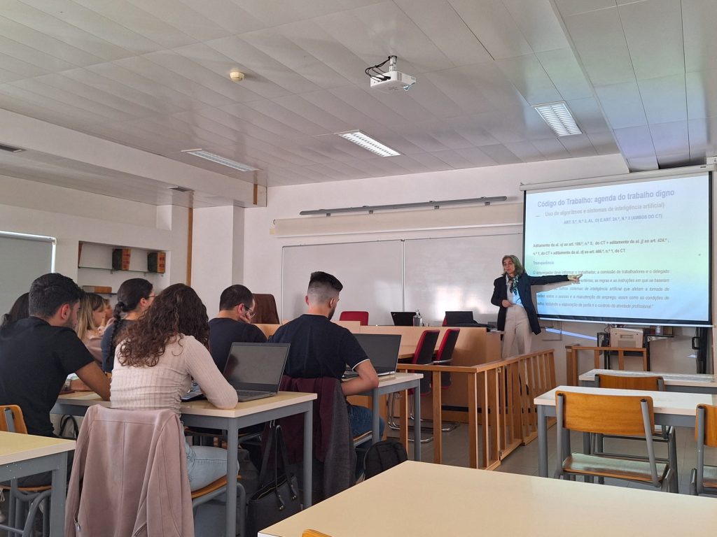 Sala de aula com estudantes sentados nas mesas com computadores. Ao fundo, junto ao quadro, uma mulher vestida com casaco azul, calças de corcreme e um lenço ao pescoço, aponta para uma projeção com texto.