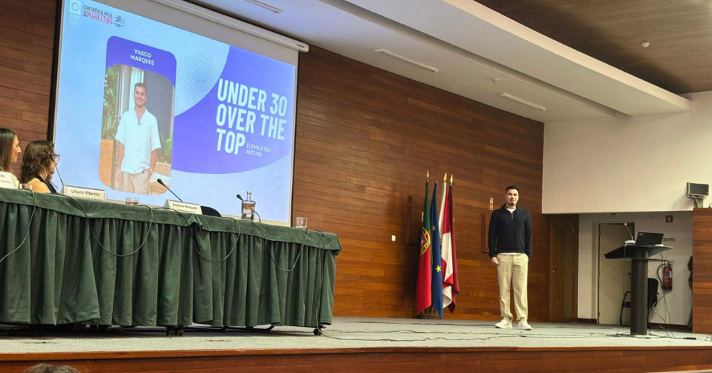 Um jovem, à direita, está a fazer uma apresentação perante uma plateia, na Conferência Anual de Marketing. À esquerda na imagem está a projeção de uma apresentação, com o texto "Under 30, Over the Top" e uma fotografia com a identificação "Vasco Marques".
Sentadas a uma mesa, à esquerda, duas mulheres olham na direção da projeção e sorriem.