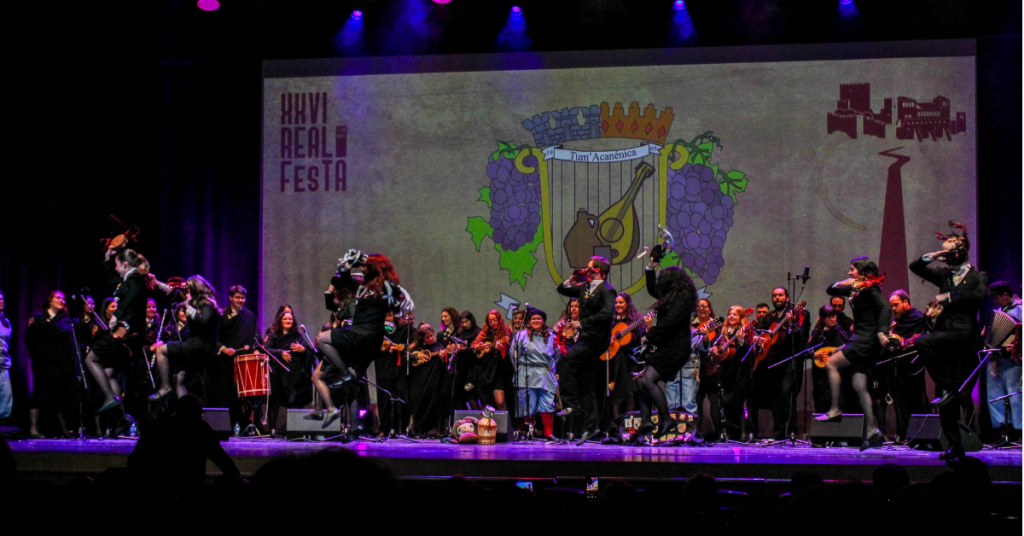 Uma tuna atua em cima de um palco durante o festiva Real FesTA. Seis participantes estão à frente e dançam com pandeireta. Atrás deles, distribuídos pelo palco, estão os restantes elementos da tuna, que cantam e tocam instrumentos. Atrás, numa projeção, pode ler-se "26.º Real FesTA".