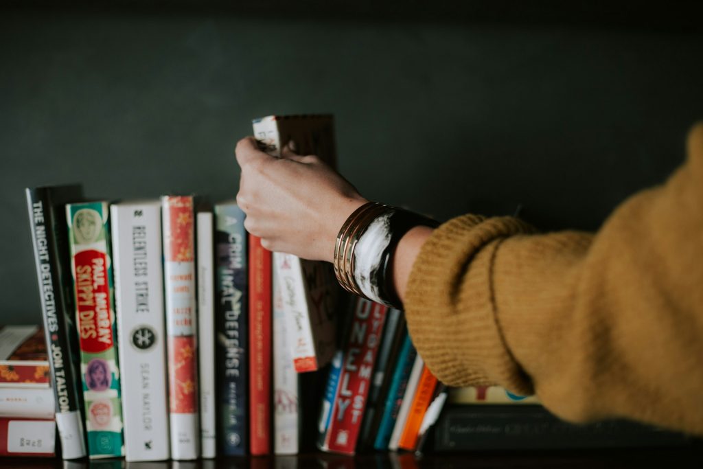 Uma mão de uma mulher estende-se em direção a uma prateleira cheia de livros na vertical, encostados uns aos outros.