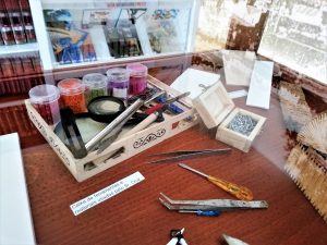 Caixa de ferramentas e materiais usados pelo artesão Luís Cruz. Dentro da caixa, pequenos frascos com missangas coloridas. De fora, uma pinça, uma lupa e um medidor.