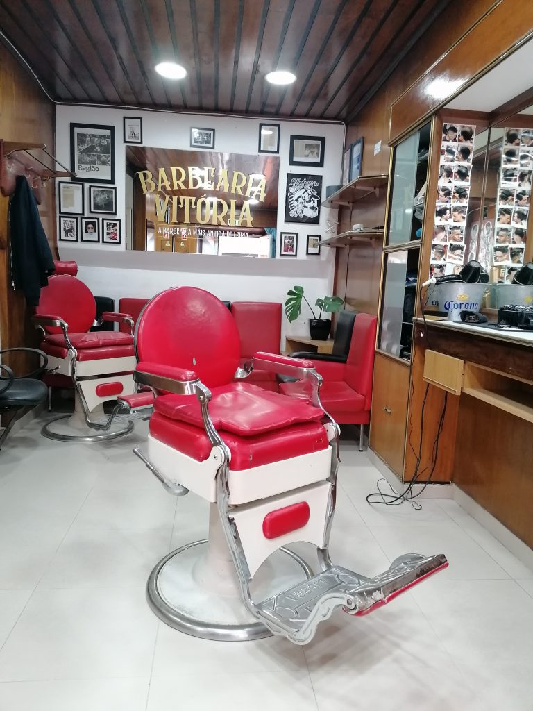 Em primeiro plano, cadeira vermelha antiga de barbeiro da Barbearia Vitória. Ao fundo, uma parede decorada com molduras de fotografias antigas e, centrado na parede, um espelho com palavras em dourado que leem "Barbearia Vitória, a barbearia mais antiga de Leiria".