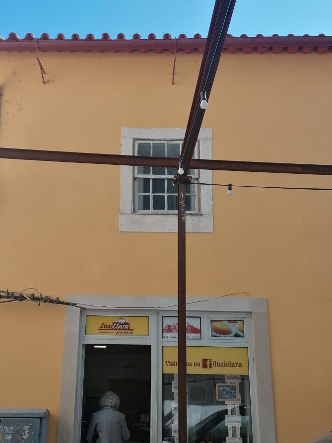 Fachada da pastelaria LuziClara. O prédio, amarelo, tem uma janela e no primeiro andar e, em baixo, a porta de entrada da loja, por onde entra uma cliente.