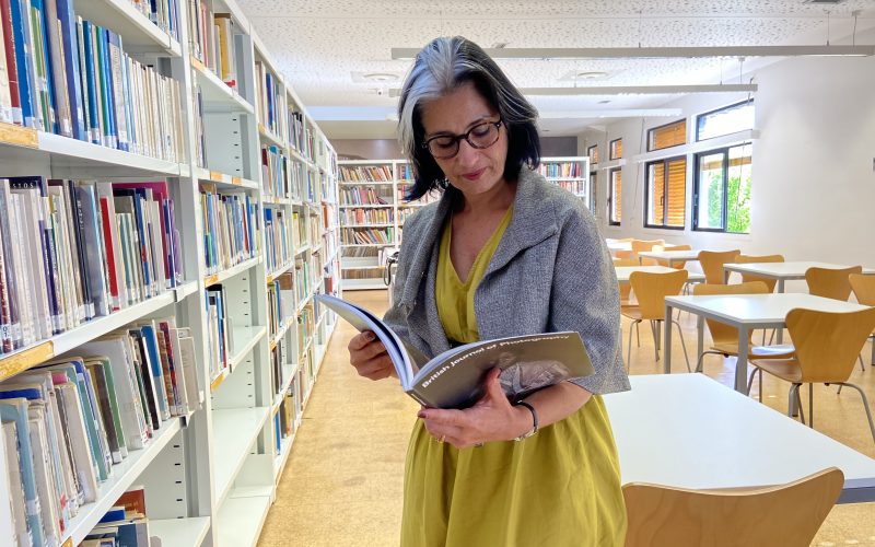 Sara Brighenti está numa biblioteca. Olha para um livro aberto que tem nas mãos. Veste um vestido verde e um casaco cinzento. Do lado esquerdo e atrás de si estão estantes cheias de livros e mesas e cadeiras.