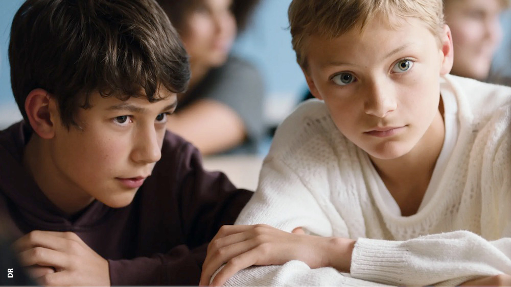 Frame do filme Close. Dois meninos com os braços cruzandos em cima de uma mesa em sala de aula.
