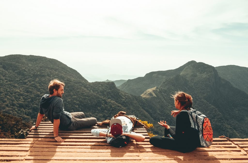 Três jovens a falar, sentados num miradouro, com vista para as montanhas. Cenário de férias, com sol.
