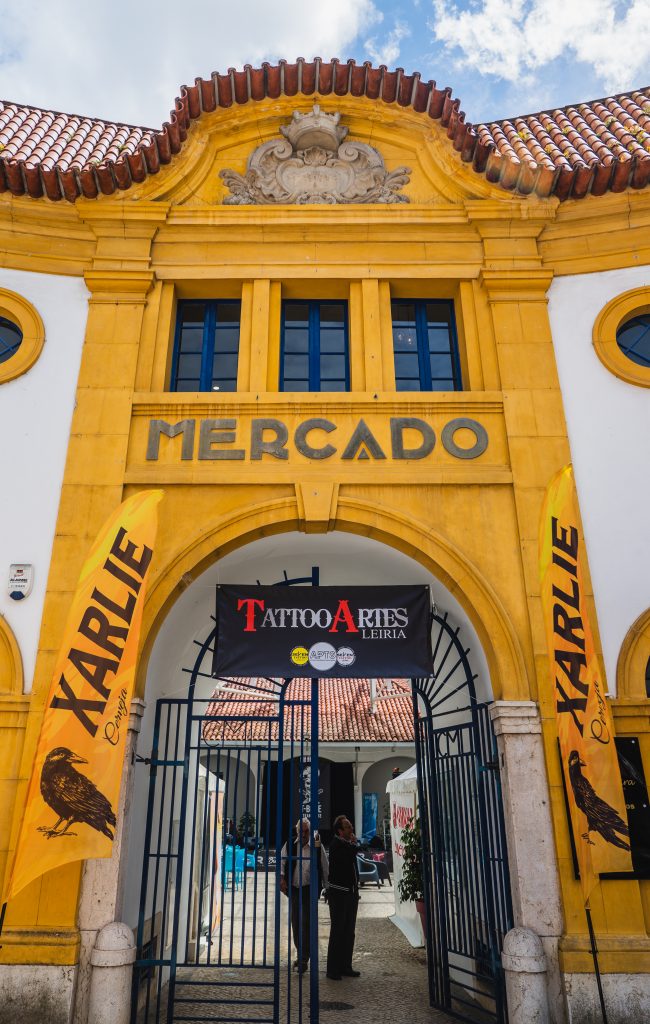 Fachada do Mercado de Sant'Ana, em Leiria, espaço onde se realizou o evento TattooArtes. A entrada tem um portão de ferro azul e o edifício é amarelo e branco. Há três pequenas janelas com caixilhos de cor azul. Na fachada está escrito em letras maiúsculas: Mercado.