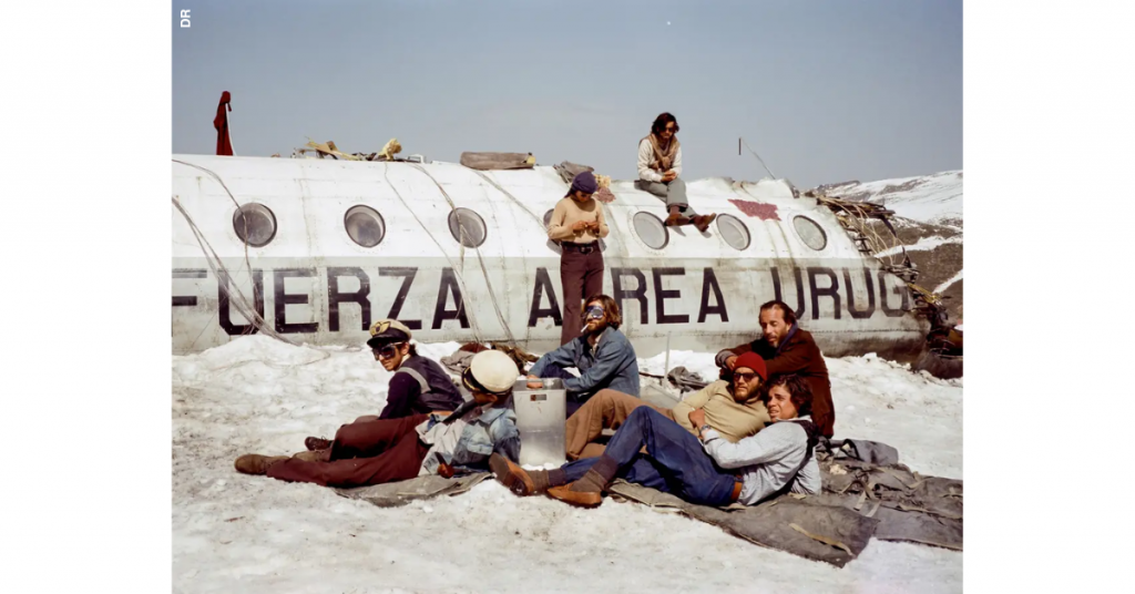 É uma imagem da série A Sociedade da Neve. Mostra seis pessoas deitadas na neve, em cima de mantas, à frente do destroço de um avião em cuja lateral se lê "Fuerza Aerea Uruguaya". Em pé, encostado à carcaça do avião está um homem e sentado em cima do avião, outro.