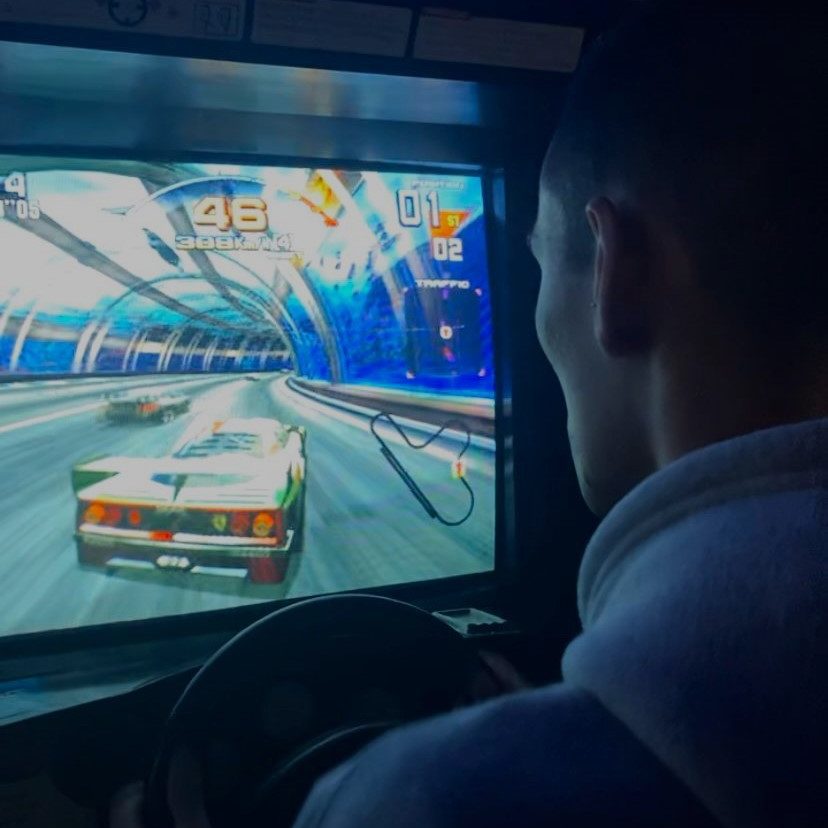 Rapaz, visto de traz, joga um jogo de carros, que conduz numa máquina de arcade. Vemos o monitor de frente, onde aparece um carro.