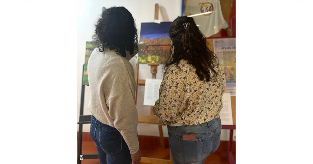 Duas mulheres são vistas de costas a olhar em direção a uma tela com um pintura de várias cores, que se vê ao fundo. Trata-se de uma das peças que integra a exposição "Asas de Abril: Voando com as cores e as palavras" realizada pelos estudantes do Programa 60+ do Politécnico de Leiria.