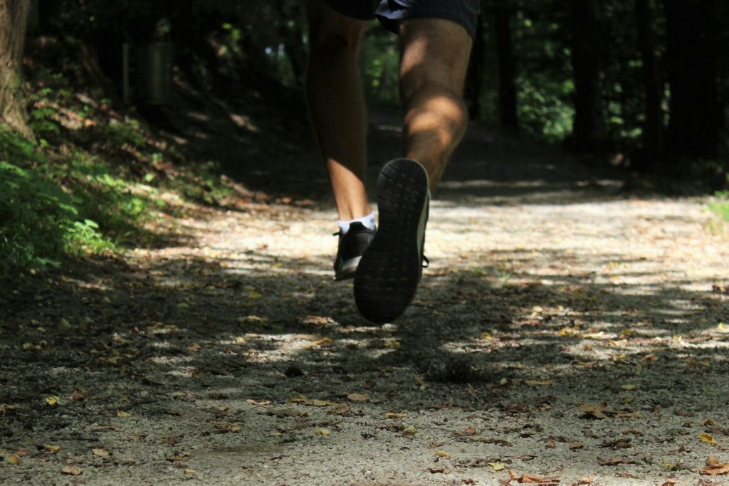 A foto mostra uma pessoa a correr, vista de trás, dos joelhos para baixo. Usa calções e sapatilhas pretas. O percurso é um trail de mata.