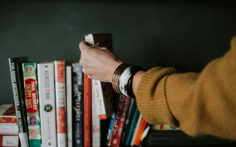 Uma mão de uma mulher estende-se em direção a uma prateleira cheia de livros na vertical, encostados uns aos outros.